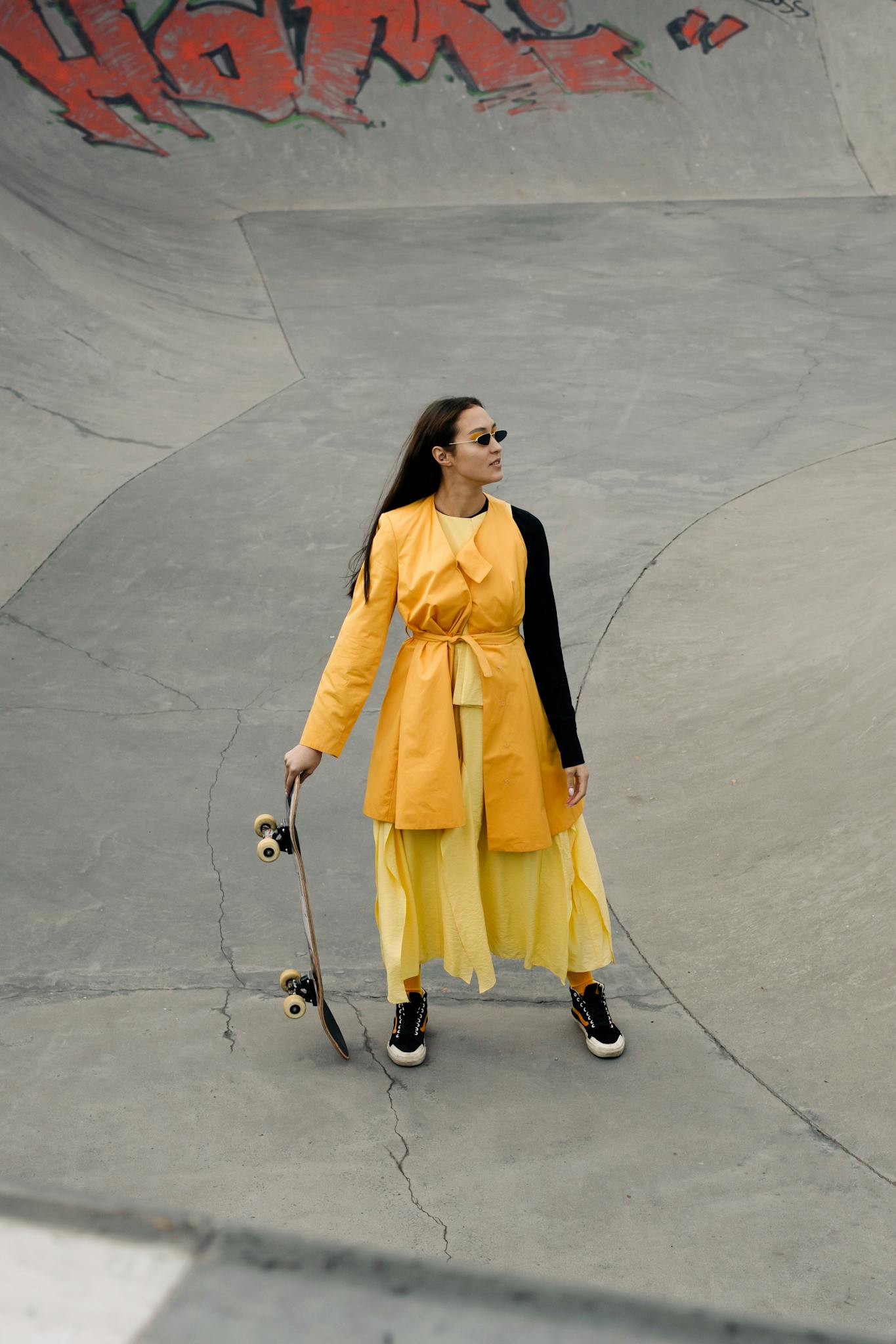 Stylish woman in a yellow dress with skateboard in a concrete skate park, exuding confidence and fashion.