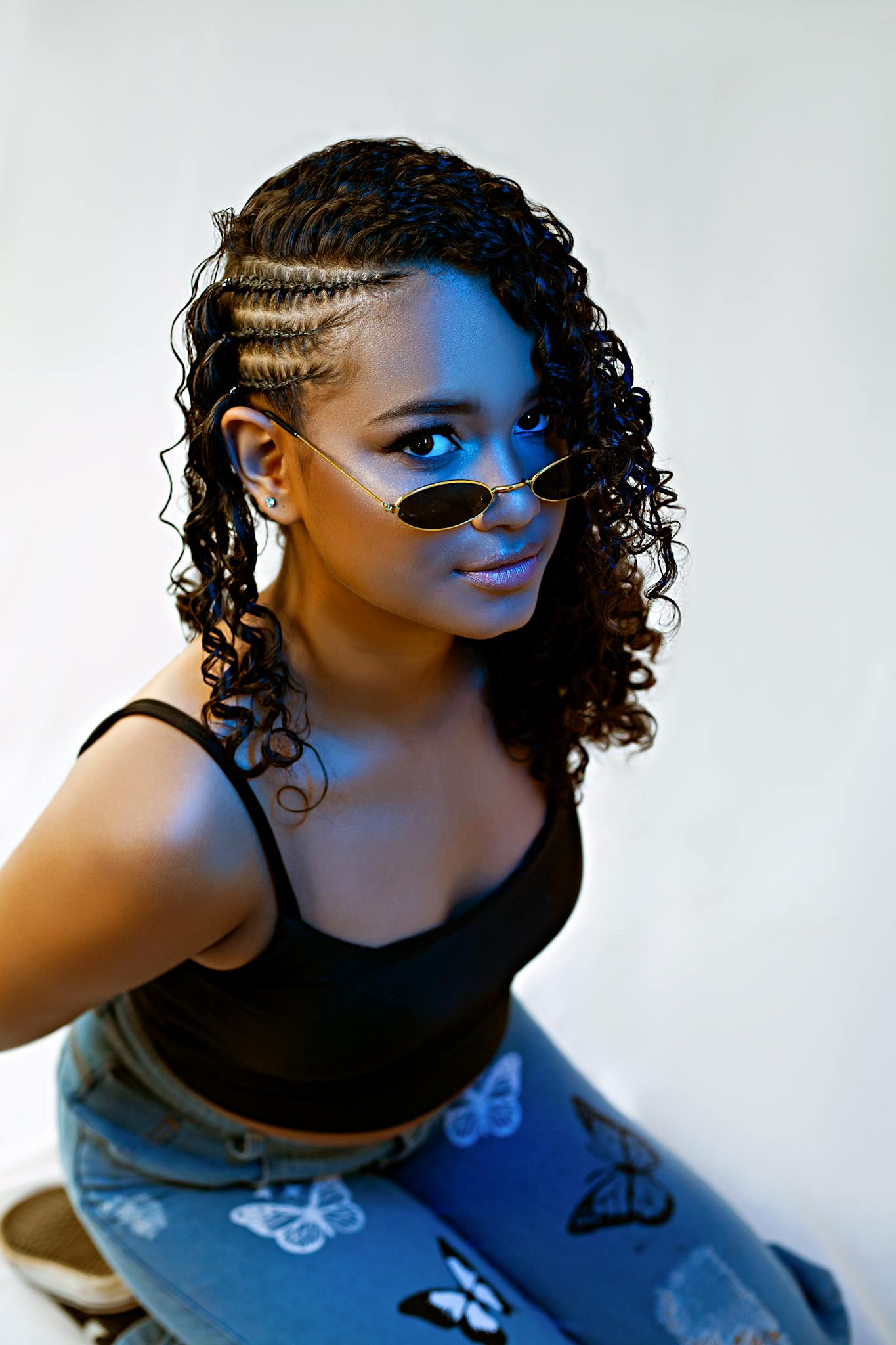 Young woman with braids posing confidently in a studio setting, wearing trendy jeans and sunglasses.